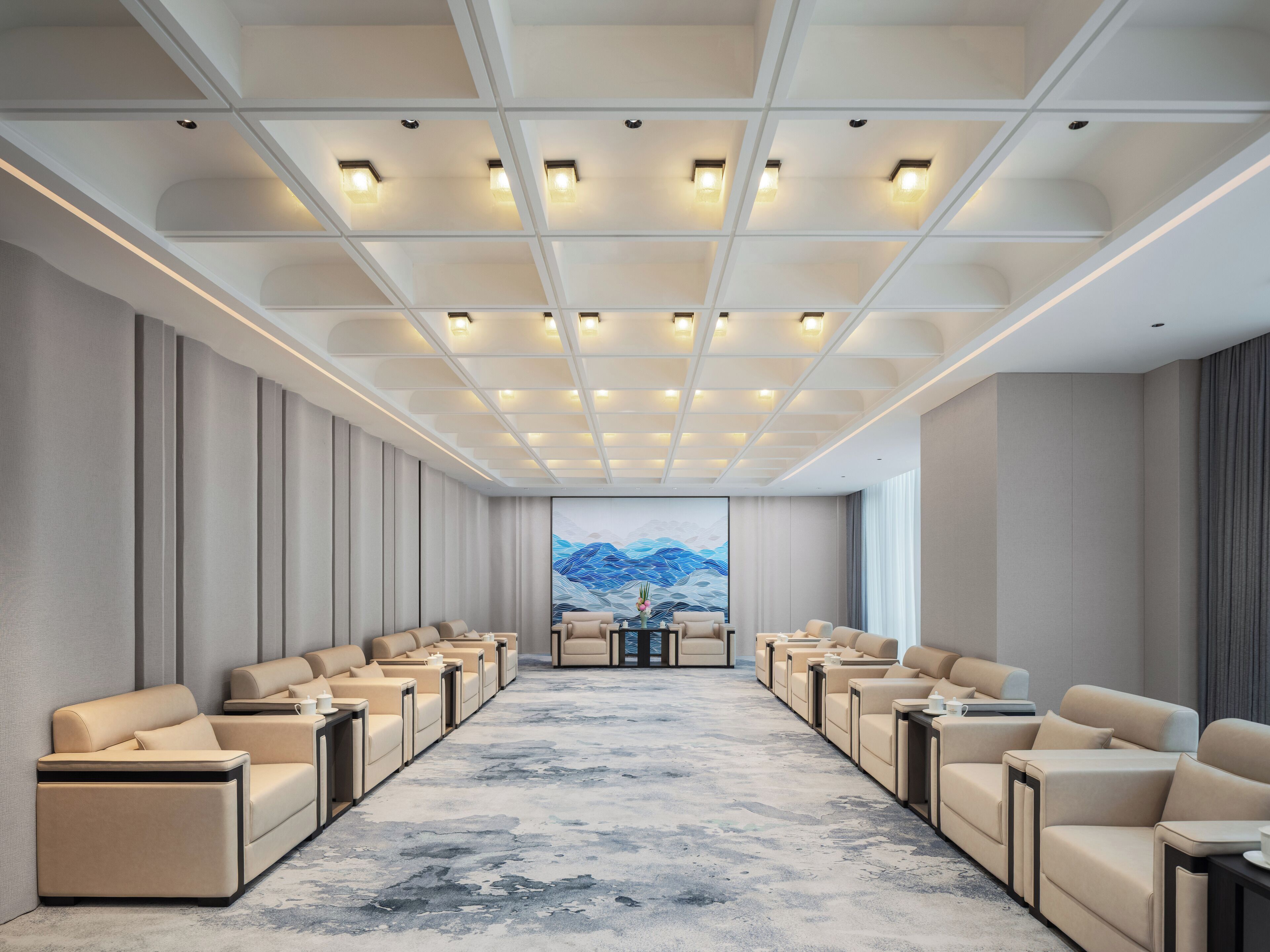 A conference room with a long row of beige sofas facing a large table, a ceiling with recessed lighting, and a painting of mountains on the far wall.