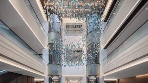 A spacious lobby with a modern design, featuring a central elevator surrounded by glass walls and a decorative ceiling installation. - ADEN Hotel Shenzhen Nanshan (Shenzhen)