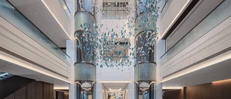 A spacious lobby with a modern design, featuring a central elevator surrounded by glass walls and a decorative ceiling installation.