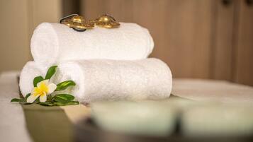 Two rolled white towels with a pair of golden bells on top, placed on a white surface with a green plant and a yellow flower beside them.