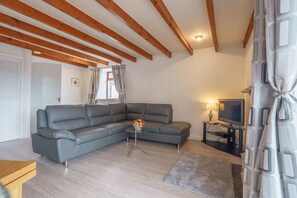 Ein modernes Wohnzimmer mit einem grauen Ecksofa, einem Glastisch, einem Flachbildfernseher auf einem Standfuß und einer Holzbalkendecke.