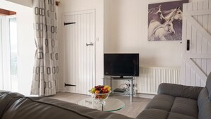 A living room with a sofa, a glass table with fruit, a television, and a door with a curtain. - Jane Welsh Cottage - a cottage that sleeps 4 guests  in 2 bedrooms (Thornhill)