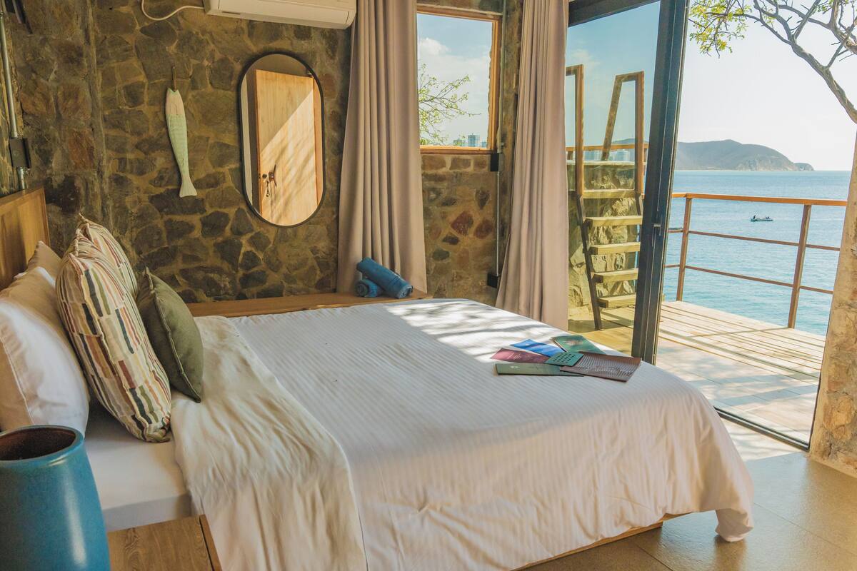 A bedroom with a stone wall, a wooden bed, a window with a view of the sea, and a balcony with a railing.