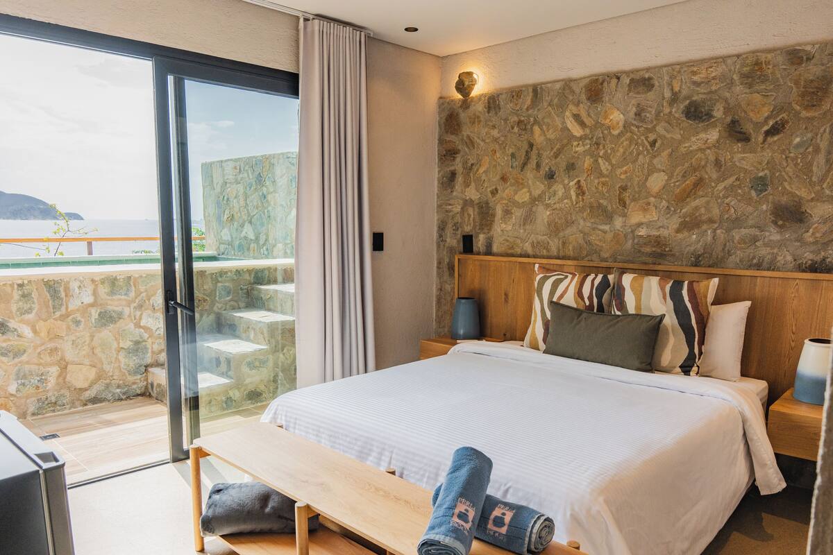 A bedroom with a stone wall, a bed with multiple pillows, a bench with towels, and a view of the sea through a glass door.