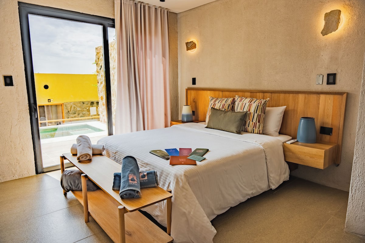 A hotel room with a bed, bedside tables, a bench, a view of the outside, and a small table with towels and a camera.