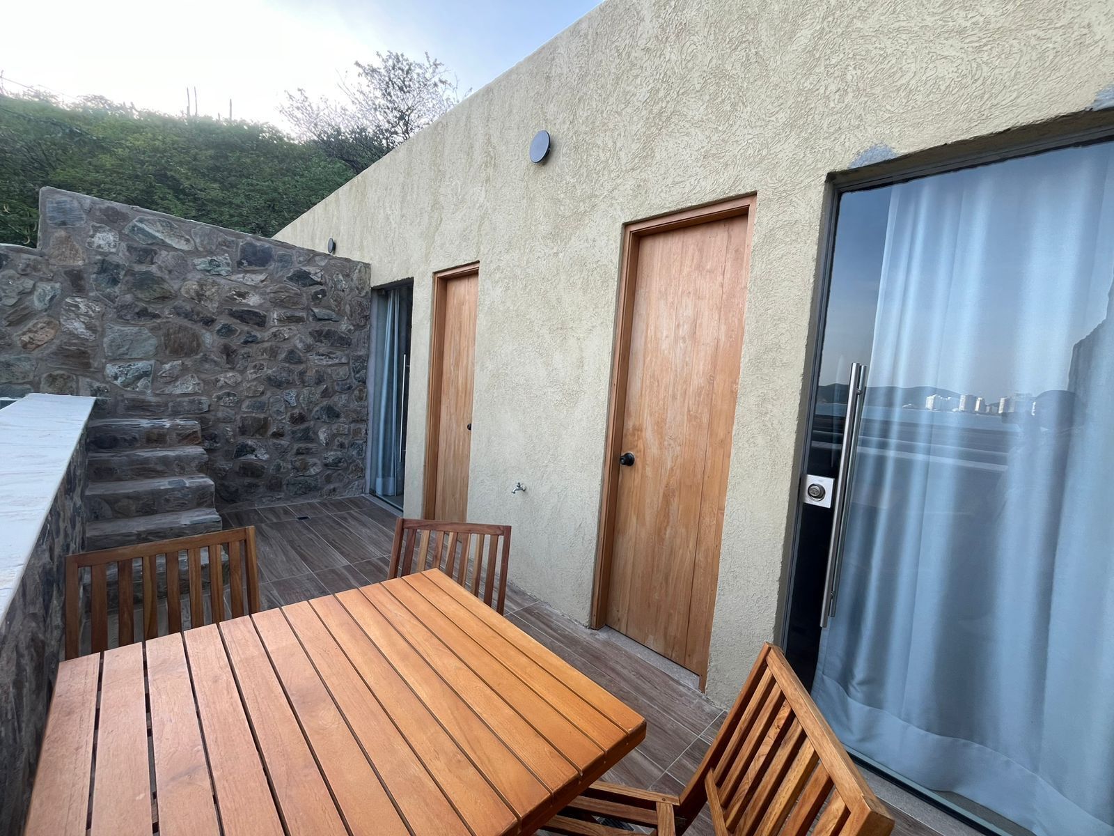 A patio with a wooden table and chairs, a stone wall, and a door leading to a covered area.