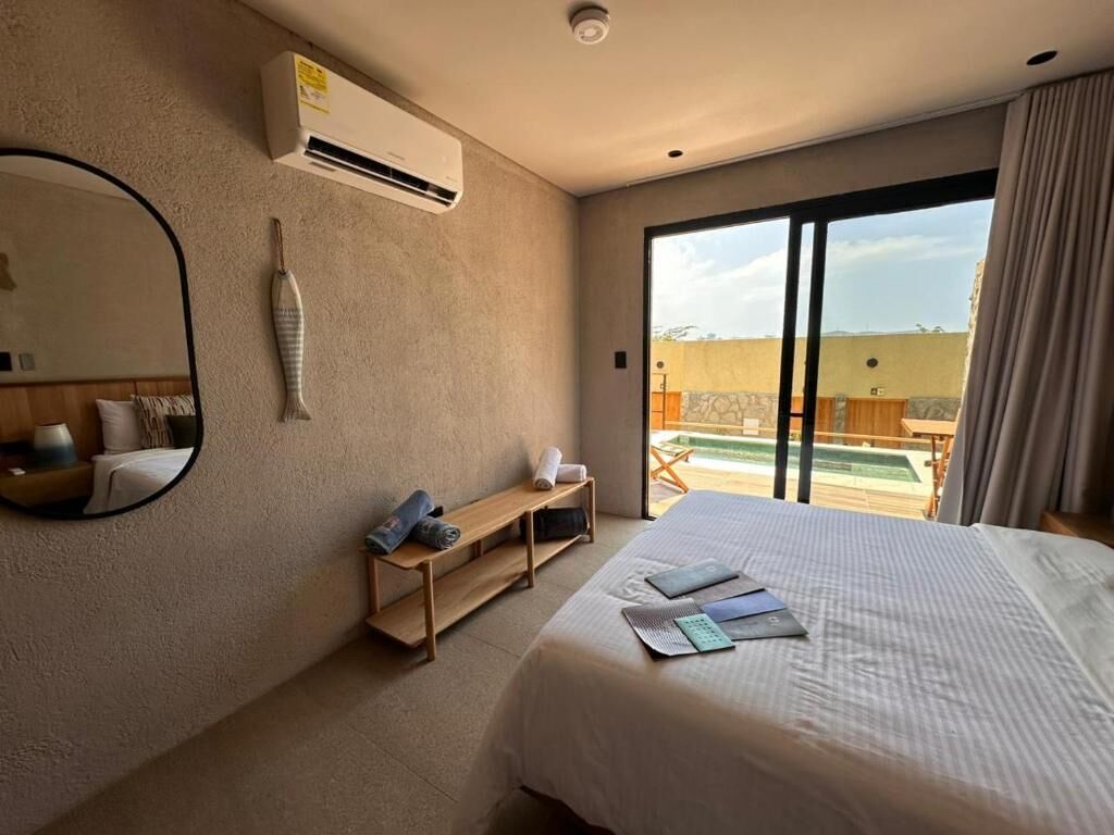 A hotel room with a bed, a mirror, a wooden shelf, an air conditioning unit, and a view of the outside.