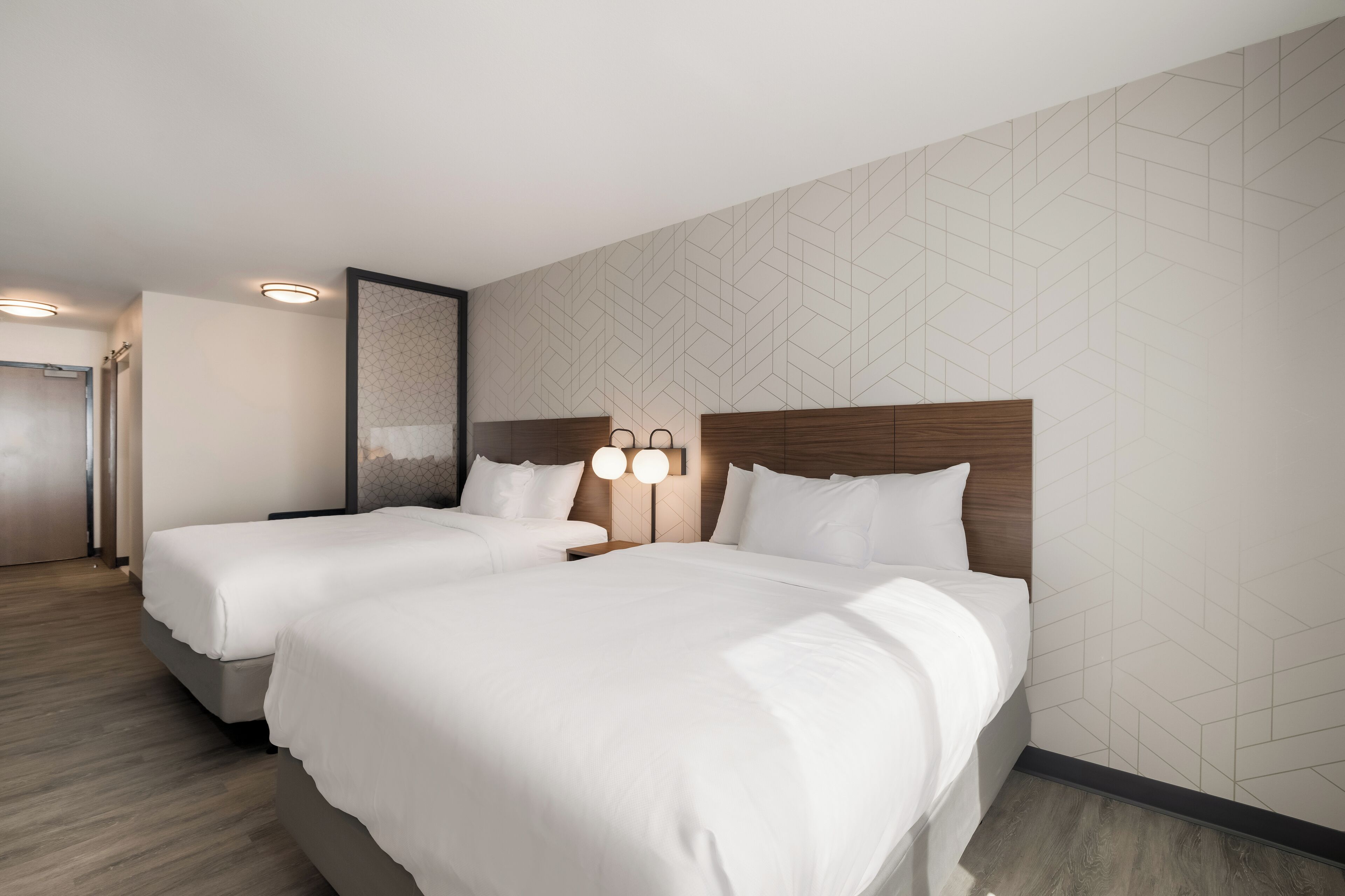 Une chambre d’hôtel avec deux lits, une tête de lit en bois, un luminaire fixé au mur et un mur orné d’un motif géométrique.