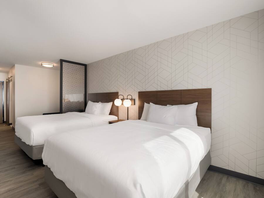 Une chambre d’hôtel avec deux lits, une tête de lit en bois, un luminaire fixé au mur et un mur orné d’un motif géométrique.