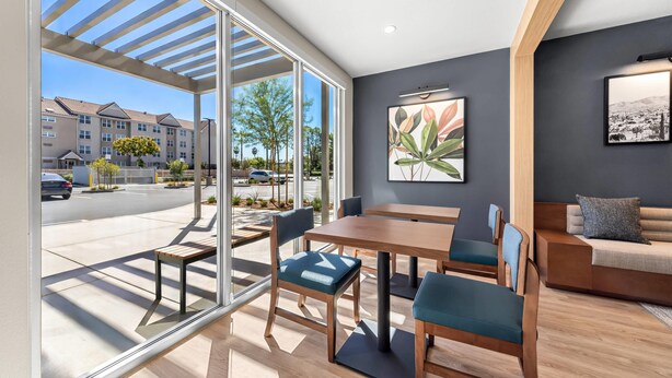 A modern interior with a dining area, a sofa, and a large window offering a view of the street. - Everhome Suites San Bernardino - Loma Linda (San Bernardino)