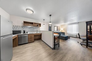 A modern kitchen with a built-in oven, microwave, and coffee maker. There is a dining area with a table and chairs, and a sofa. The room has wooden flooring and a large window with blinds. - Everhome Suites San Bernardino - Loma Linda (San Bernardino)