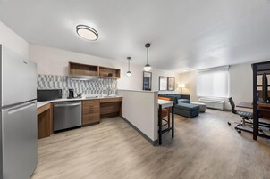 Cocina moderna con horno empotrado, microondas y cafetera. Cuenta con un comedor con mesa y sillas, además de un sofá. El ambiente tiene suelo de madera y un ventanal con persianas.