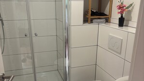 A bathroom with a glass-enclosed shower, a toilet, a sink, and a shelf with towels and a vase. - Cozy apartment directly on the Rhine (Wiesbaden)