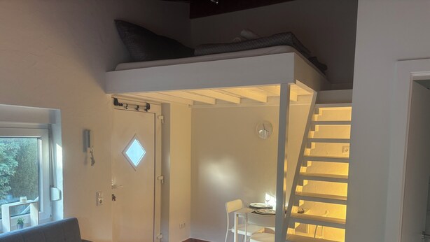 A compact living space with a sofa, a dining area with chairs and a table, a staircase, and a door with a window. - Cozy apartment directly on the Rhine (Wiesbaden)