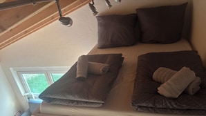A bed with dark bedding and two rolled towels, a window with a view of greenery, and a wooden ceiling. - Cozy apartment directly on the Rhine (Wiesbaden)