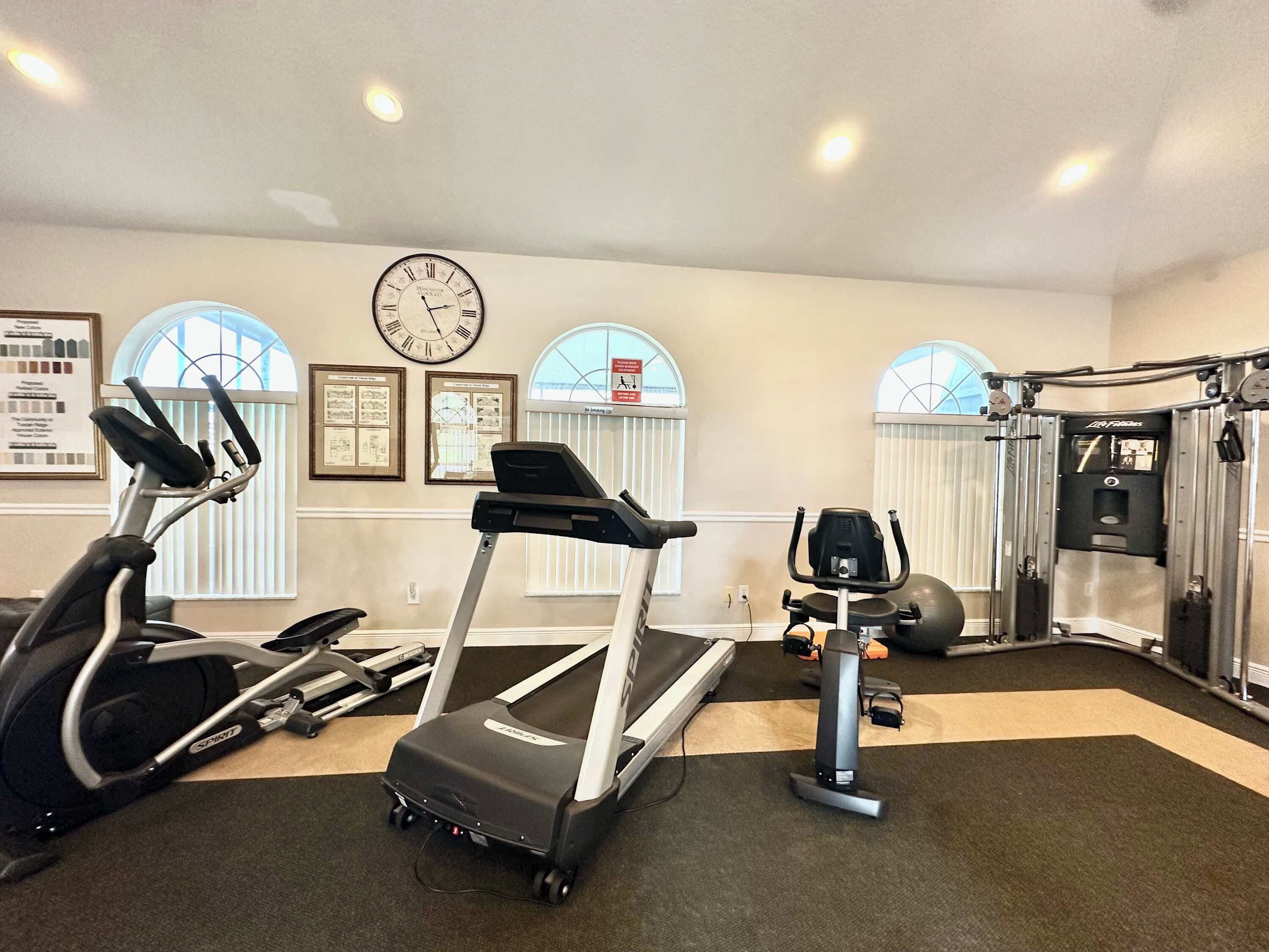 Un gimnasio con cintas de correr, bicicletas estáticas y máquinas de pesas. Hay un reloj en la pared y ventanas grandes con persianas.