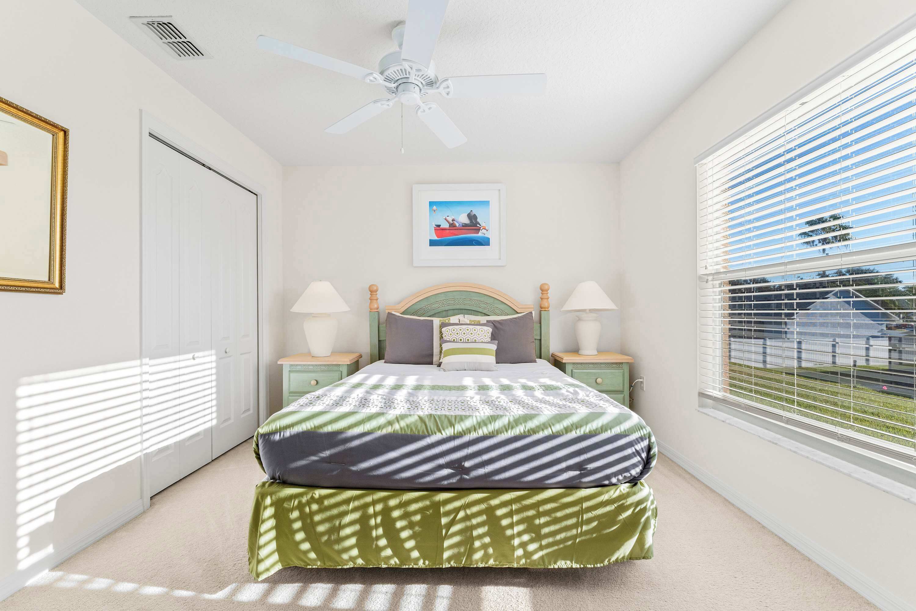A bedroom with a bed, a ceiling fan, a window with blinds, a framed picture on the wall, and a door.