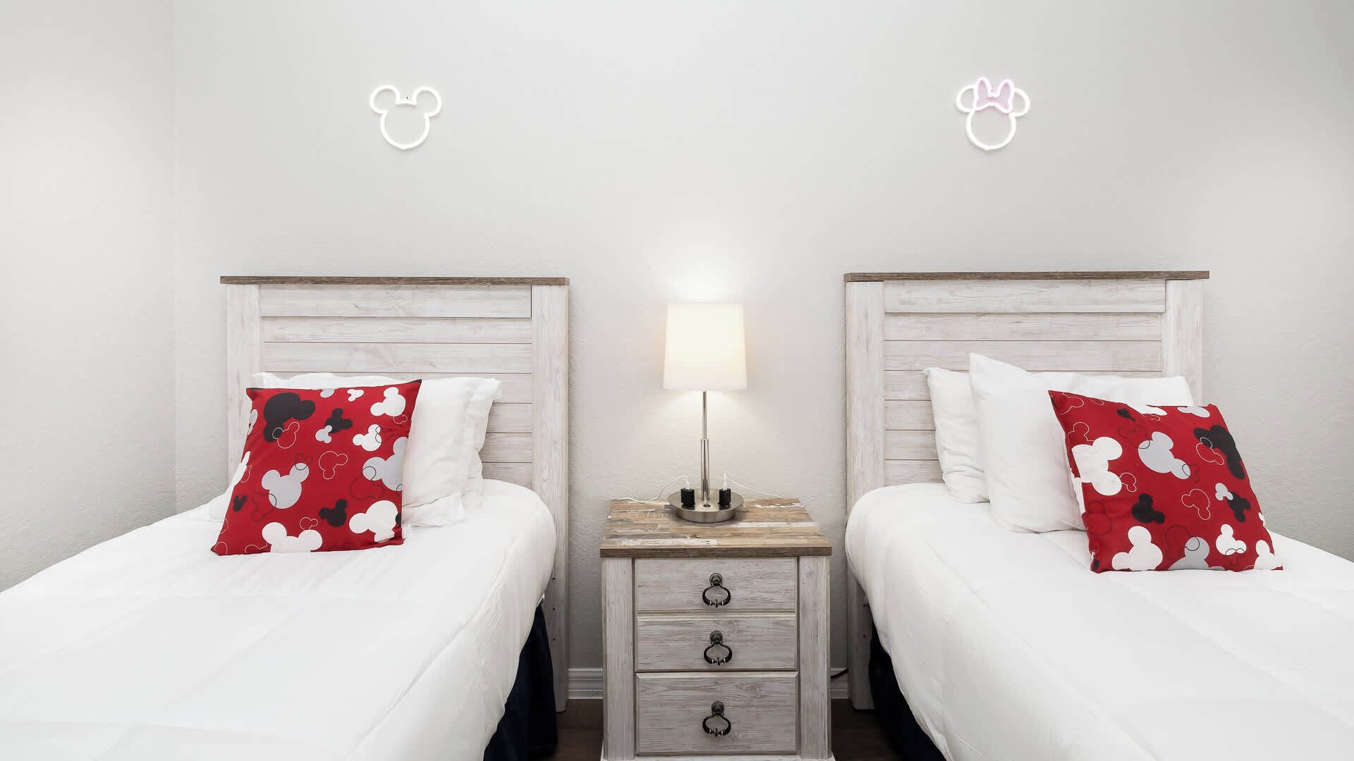 Une chambre d’hôtel avec deux lits, chacun recouvert d’une courtepointe blanche et agrémenté d’oreillers rouges ornés d’un motif de personnage de dessin animé. Une table de chevet avec une lampe se trouve entre les lits, et un meuble à tiroirs en bois est placé en dessous.
