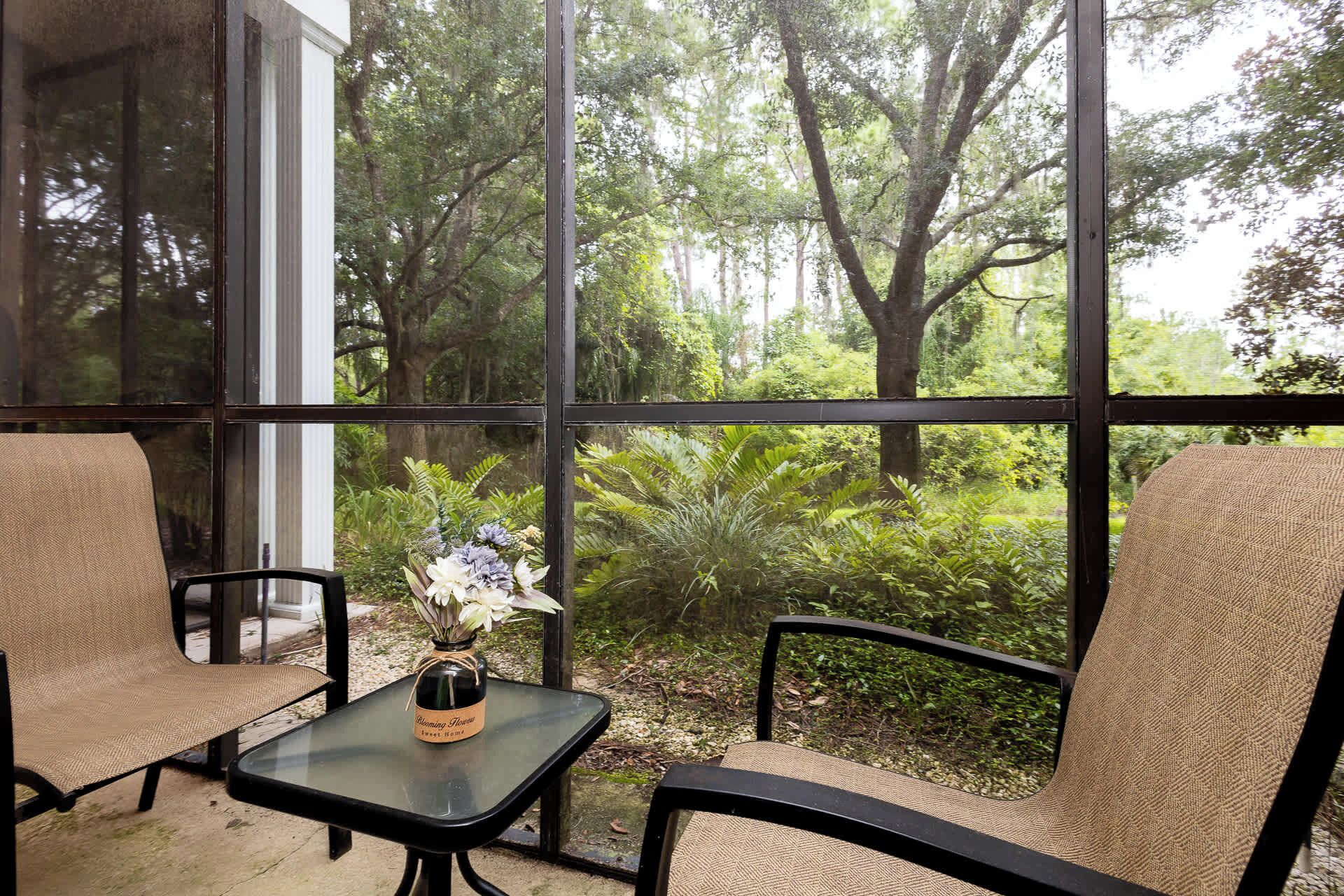 A conservatory with two wicker chairs and a glass table, featuring a view of a lush garden through large windows.