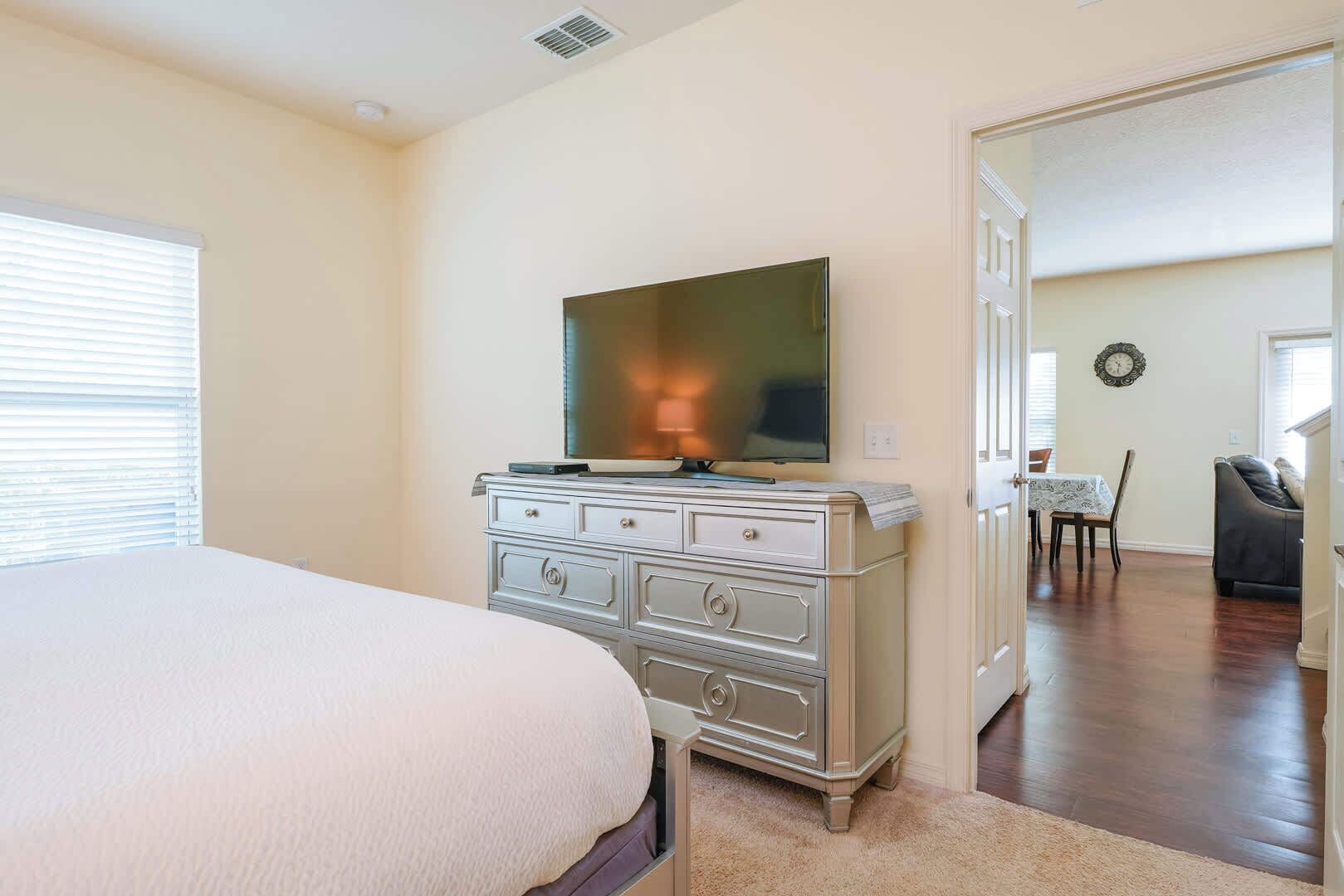 A bedroom with a bed, a dresser, a television, and a dining area visible through an open door.