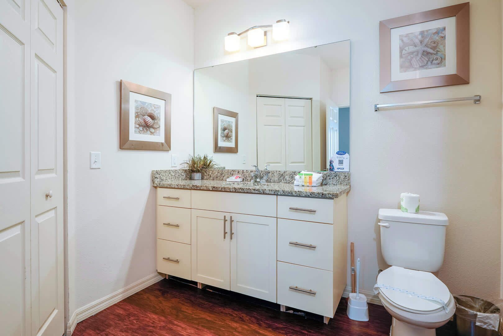 Une salle de bain avec des toilettes blanches, un lavabo doté d’un plan de travail en granit, un miroir au-dessus du lavabo et un tableau encadré au mur.