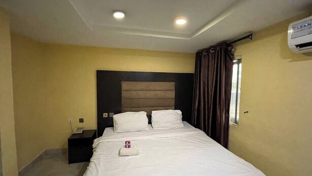 A hotel room with a bed, a nightstand, a window with curtains, and an air conditioning unit. - suite 104 (Enugu)