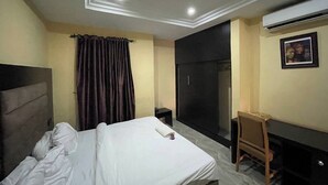 A hotel room with a bed, a desk, a chair, a wardrobe, and an air conditioning unit. - suite 104 (Enugu)
