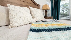 Una cama con colcha y dos almohadas blancas, cabecera de madera, una lámpara y una ventana con cortinas.