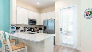 A modern kitchen with white cabinets, a stainless steel refrigerator, a microwave, and a coffee maker. There are two white chairs with woven seats.