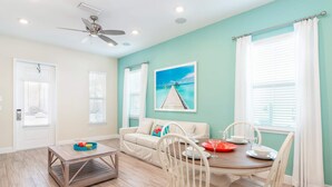 A bright living room with a teal accent wall, a white sofa, a wooden coffee table, and a dining area with white chairs and a wooden table.