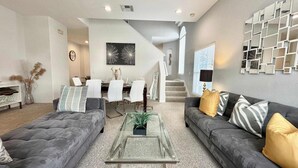 A modern living room with two grey sofas, a glass coffee table, and a dining area with white chairs.