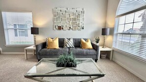 A living room with a grey sofa, two side tables, a glass coffee table, and a decorative wall piece.