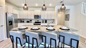 A modern kitchen with white cabinets, a stainless steel refrigerator, a microwave, and a double oven. There are four bar stools around a granite island with a sink and a faucet. - Adventure of a Lifetime! 5 BDR at Windsor Island (Davenport)