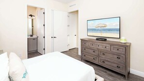 Habitación de hotel con cama, cómoda, televisor mostrando una escena de playa y baño visible a través de una puerta abierta.