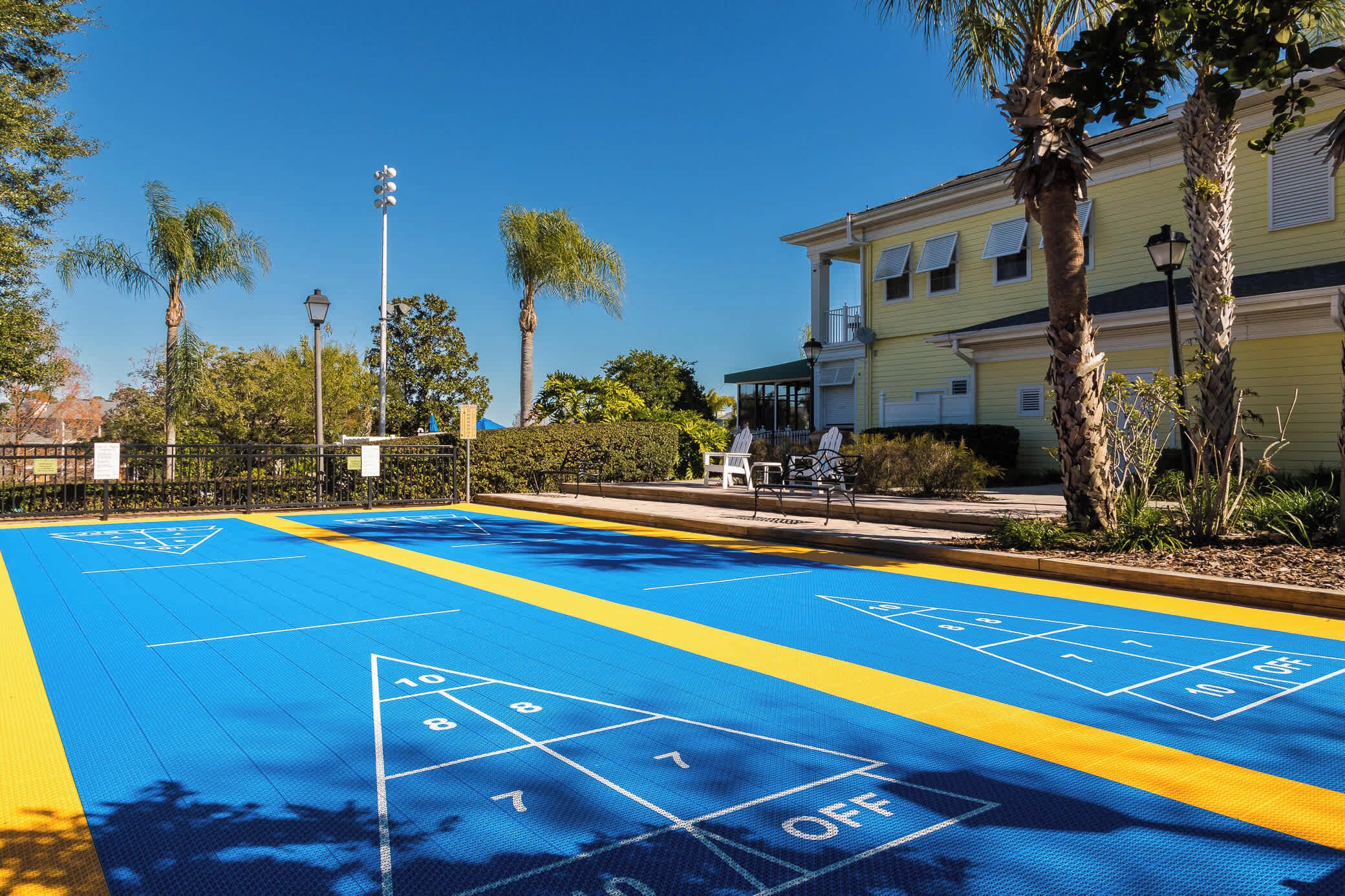 Un campo da shuffleboard con superficie blu e gialla, circondato da palme e un edificio con un balcone.