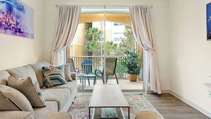 A living room with a sofa, coffee table, and a view of a balcony with plants and a building in the background.