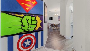 A hallway with a Superman and Captain America mural, a dining area with a TV, and a kitchen in the background.