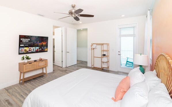 A bedroom with a large bed, a TV mounted on the wall, a ceiling fan, and a door leading to a balcony.