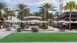 An outdoor seating area with tables and umbrellas, surrounded by palm trees and a building with a sign. - Up to Scale With Margaritas (Kissimmee)