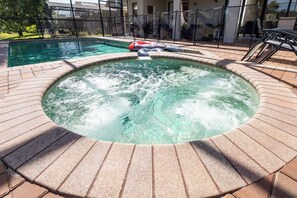 A hot tub with a circular brick surround, a pool in the background, and a fenced area with chairs.