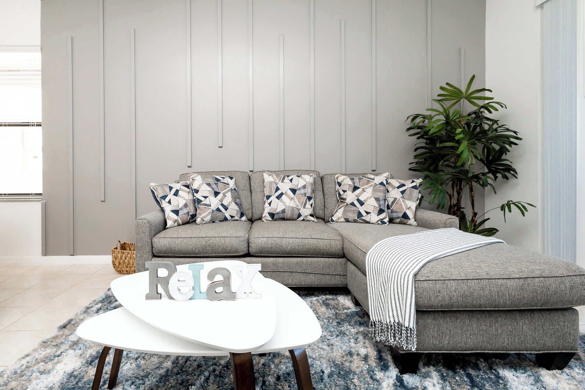 Una sala de estar moderna con un sofá seccional gris, almohadones estampados, una mesa de centro blanca y una planta grande.