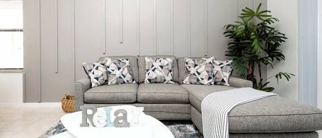 Una sala de estar moderna con un sofá seccional gris, almohadones estampados, una mesa de centro blanca y una planta grande.