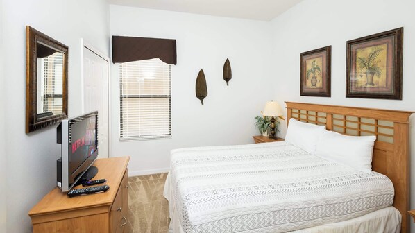 A hotel room with a bed, a TV on a wooden nightstand, a window with blinds, and two wall-mounted pictures.