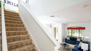 Un escalier mène à un espace de vie comprenant un canapé bleu, une petite table et une fenêtre avec des stores.