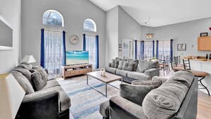 Una sala de estar amplia con un televisor grande, sofás grises, una mesa de centro y un comedor con una cocina al fondo.