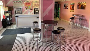 Ein Café im Retro-Stil mit Rosa- und Weißtönen, das eine Verkaufstheke mit Softeis-Kegeln und -Bechern, einen Barbereich mit Hockern und einem gemusterten Boden zeigt.