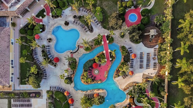 Eine Vogelperspektive zeigt ein Resort mit mehreren Schwimmbecken, Liegestühlen und Palmen.