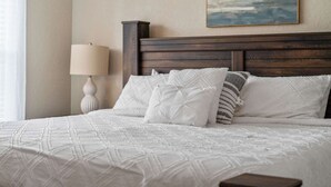 Ein ordentlich bezogenes Bett mit weißer Decke und Kissen, ein Holz-Kopfteil, eine Nachttischlampe und ein Gemälde an der Wand.