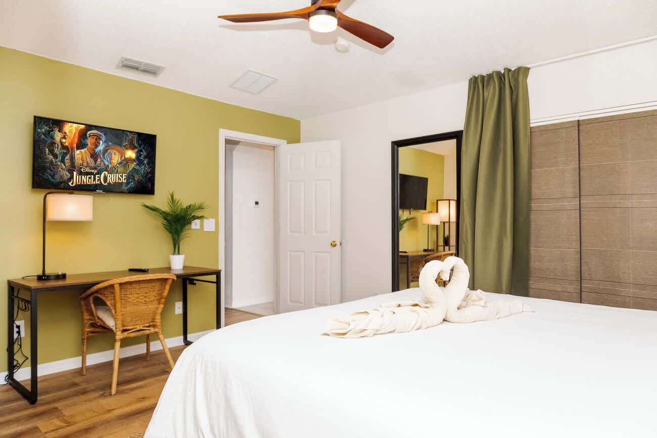 A hotel room with a bed, a desk, a chair, a ceiling fan, a wall-mounted TV, and a door.