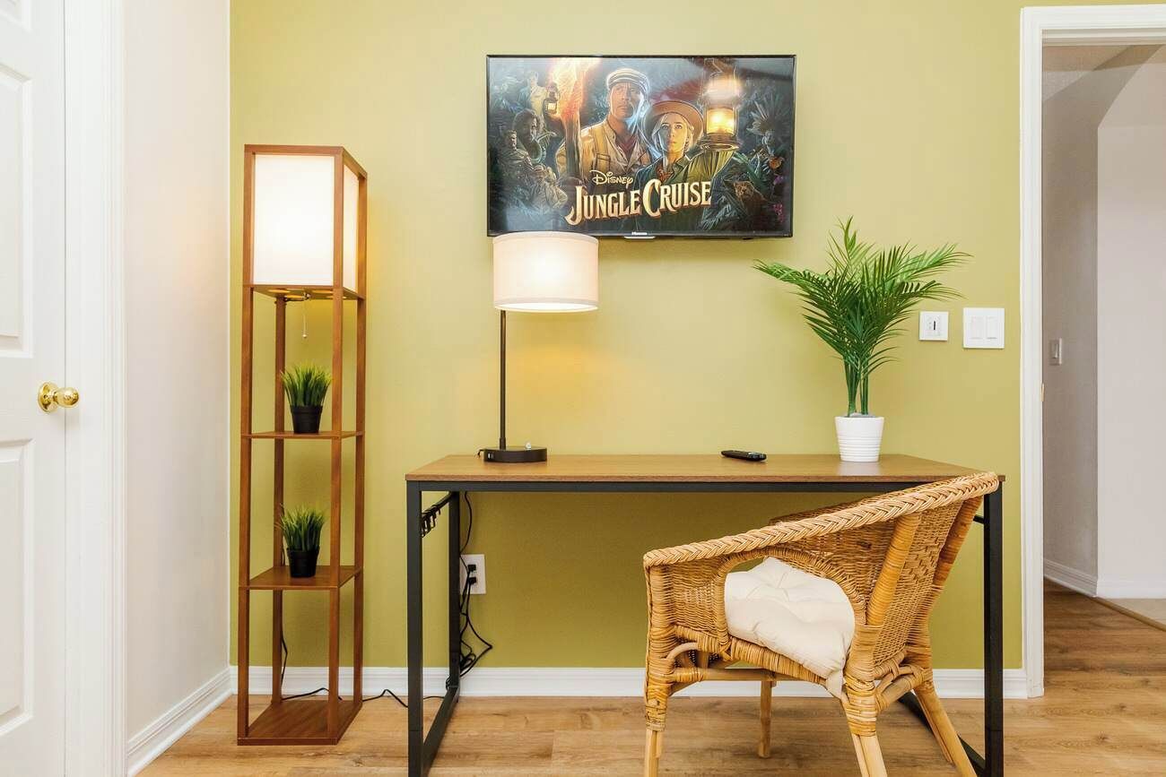 Une pièce comprenant un bureau, une chaise, un téléviseur fixé au mur diffusant le film « Disney Jungle Cruise », une lampe sur pied et deux plantes en pot.